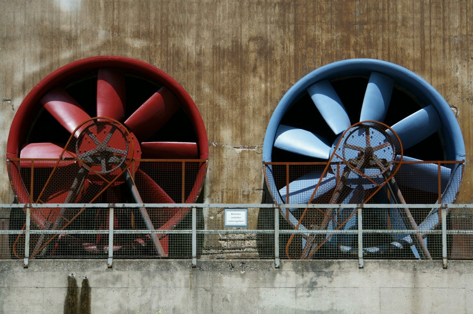Photo by Pixabay - Czyste Powietrze - Audyty Close-up of large red and blue industrial fans on a rustic wall showcasing machinery and ventilation.